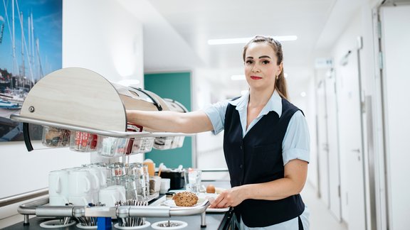 Im Krankenhausflur steht der Servicewagen, eine Service-Mitarbeitende serviert ein Frühstück.