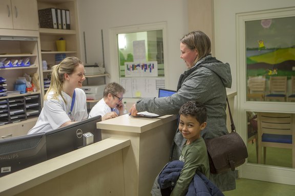 Eine Mutter mit Kind steht am Empfangstresen der Kinderstation.