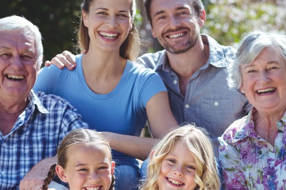 Eine fröhliche Familie bestehend aus Großeltern, Eltern und Kindern sitzen zusammen im Grünen und lächeln in die Kamera.