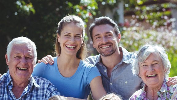 Eine fröhliche Familie bestehend aus Großeltern, Eltern und Kindern sitzen zusammen im Grünen und lächeln in die Kamera.