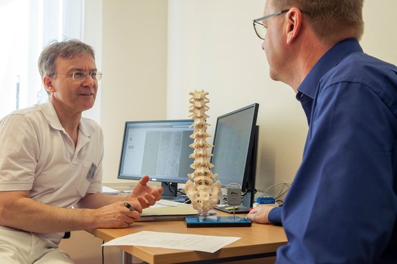 Dr. med. Wolfram Kluge klärt einen Patienten anhand eines Wirbelsäulenmodells über die SCS-Rückemankstimulation auf.