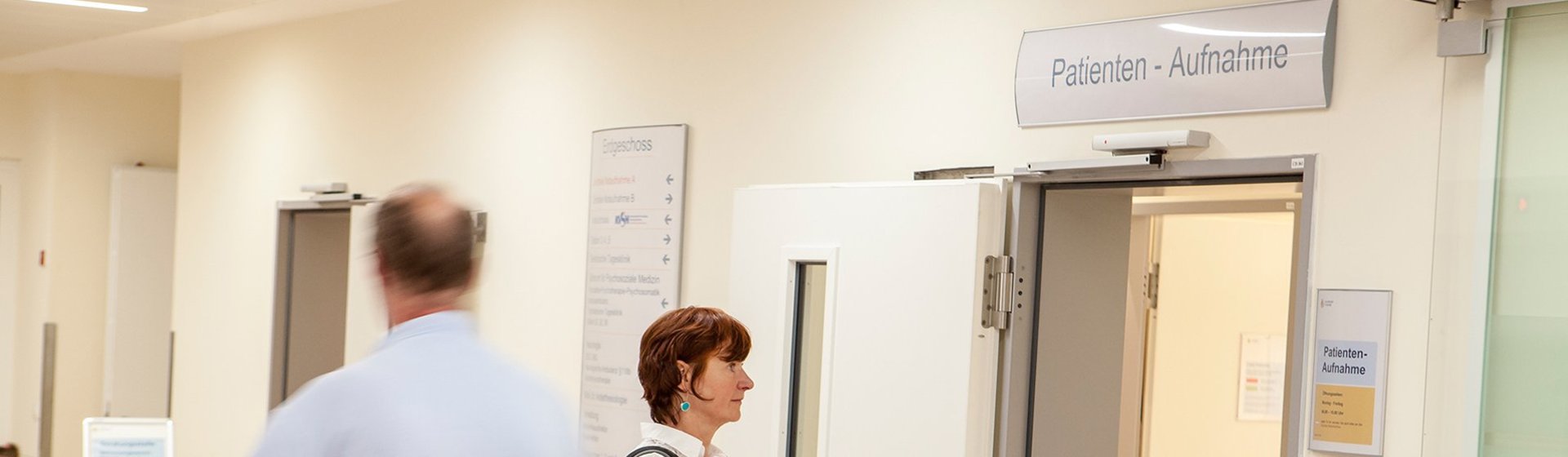 Eine Patientin geht in die Patientenaufnahme, ein Logistik-Mitarbeiter schiebt ein Patientenbett.