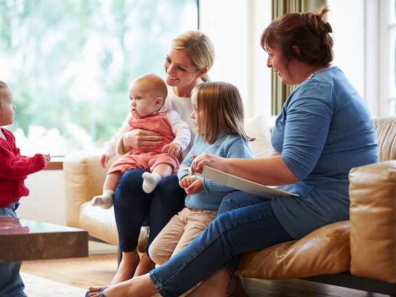 Zwei Frauen sitzen mit drei Kindern unterschiedlichen Alters in einem hellen Wohnzimmer