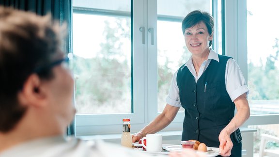 Stationsassistentin vom Service-Team serviert einem Patienten eine Mahlzeit.