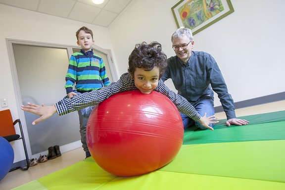 Physiotherapeut Sven Olde mit zwei Kindern in der Therapiesituation auf einer Gymnastikmatte, ein Kind liegt bäuchlings auf einem roten Gymnastikball, das zweite Kind hält die Füße des ersten Kindes.